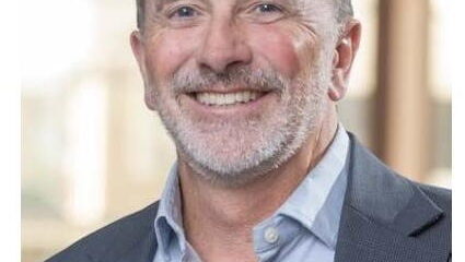 Smiling middle-aged man in a blazer and light blue shirt, wearing a name badge that reads a person’s title.